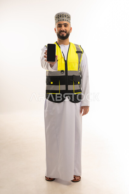 عرض شاشة سوداء فارغة عبر الهاتف المحمول، استخدام الأجهزة التقنية بالإشراف على العمل، دمج التكنولوجيا بالمجال الهندسي، مفهوم الهندسة المعمارية، بورتريه لمهندس عربي خليجي عماني يرتدي الدشداشة والكمه وسترة الحماية يمسك الجوال بيده، الجسم بالطول الكامل، خلفية بيضاء