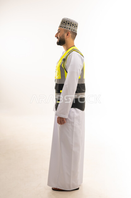 Concept of work in the engineering sector, jobs and professions for young people in the Sultanate of Oman, engineering and architectural construction, side portrait of an Arab Gulf Omani engineer wearing a dishdasha, a sleeve and a yellow protective vest looking at a place in front of him with gestures of concentration, full-length body, white background