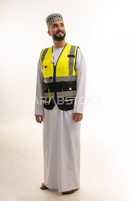 Jobs and careers for young people in the Sultanate of Oman, concept of engineering and architectural construction, work in the engineering sector, portrait of an Arab Gulf Omani engineer wearing a dishdasha, a sleeve and a yellow protective vest looking somewhere with gestures of integration and concentration, full-length body, white background