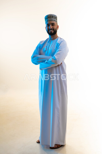 الوقوف بأيدي متقاطعة بإيماءات الثقة بالنفس، بورتريه لشاب عربي خليجي عماني يرتدي الدشداشه والكمه ينظر إلى الكاميرا بتعابير السرور والبشاشة، الاهتمام بالمظهر الخارجي والأناقة، مفهوم الرجولة والوسامة، الجسم بالطول الكامل، خلفية بيضاء