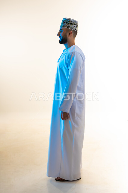 Concern for appearance and elegance, concept of masculinity and handsomeness, standing straight with gestures of self-confidence, side portrait of a young Gulf Arab Omani man wearing a dishdasha and a turban looking somewhere with expressions of pleasure and integration, full-length body, white background