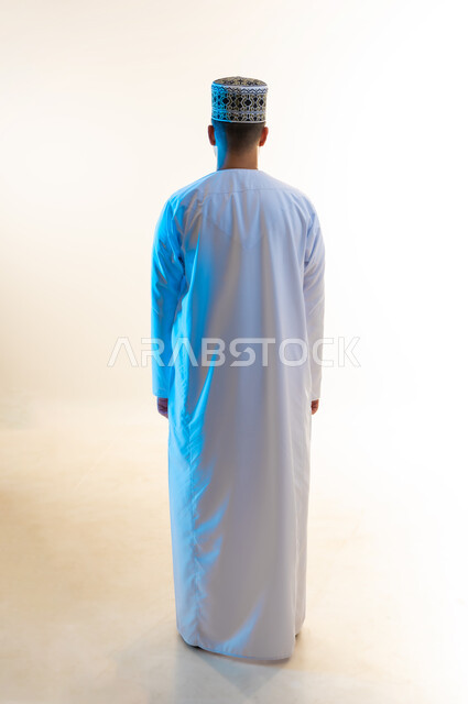 الوقوف بشكل مستقيم بإيماءات الثقة بالنفس، الاهتمام بالمظهر الخارجي والأناقة، مفهوم الرجولة والوسامة، بورتريه من الخلف لشاب عربي خليجي عماني يرتدي الدشداشه والكمه يقف بظهر مشدود، الجسم بالطول الكامل، خلفية بيضاء