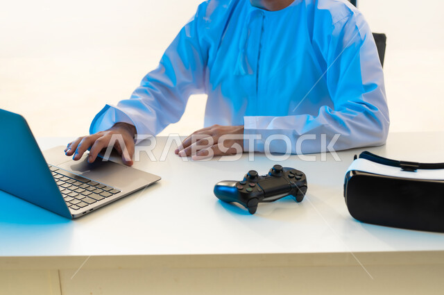 استخدام التقنيات الحديثة المتطورة، محاكاة العالم الخيالي عبر نظارات الفي آر ثلاثية الأبعاد، بورتريه مقرب لمهندس عربي خليجي عماني يرتدي الدشداشة يجلس خلف طاولة المكتب أمامه الحاسوب المحمول وبجانبه عصا التحكم بألعاب الواقع الافتراضي، خلفية بيضاء