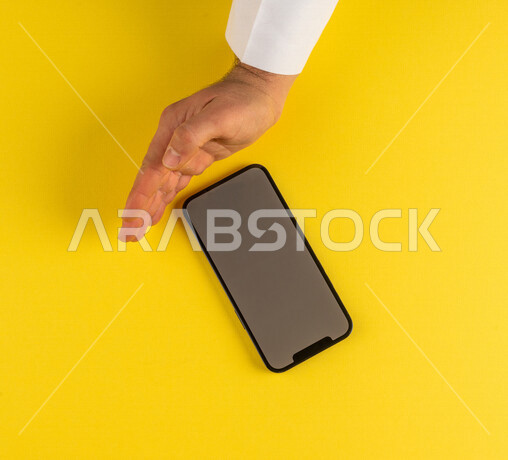 صورة مقربة ليد رجل عربي خليجي سعودي يستخدم الهاتف المحمول ، ايماءة اليد تدل على حجب الرؤيا والمشاهدة ، خلفية صفراء