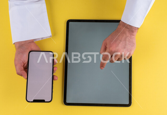 صورة مقربة ليد رجل عربي خليجي سعودي يحمل الهاتف المحمول ، ينقر على شاشة جهاز التابلت ، الترويج لموقع الويب ، التسويق الالكتروني ، تصفح المواقع الالكترونية ، استخدام الاجهزة التقنية الحديثة في الاعمال الادارية