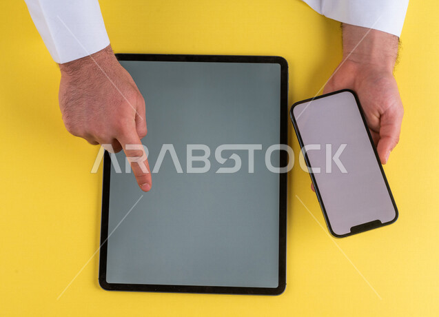 صورة مقربة ليد رجل عربي خليجي سعودي يحمل الهاتف المحمول ، ينقر على شاشة جهاز التابلت ، الترويج لموقع الويب ، التسويق الالكتروني ، تصفح المواقع الالكترونية ، استخدام الاجهزة التقنية الحديثة في الاعمال الادارية