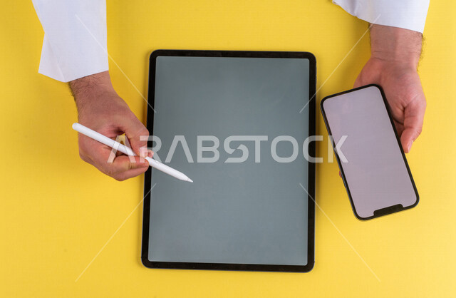 صورة مقربة ليد رجل عربي خليجي سعودي يحمل الهاتف المحمول ويستخدام جهاز التابلت ، استخدام قلم التابلت لتدوين ملاحظات خاصة بالعمل ،شرح خطوات العمل ، اعلان ترويجي لتطبيق وموقع ويب ،استخدام التقنيات الحديثة في الاعمال الادارية