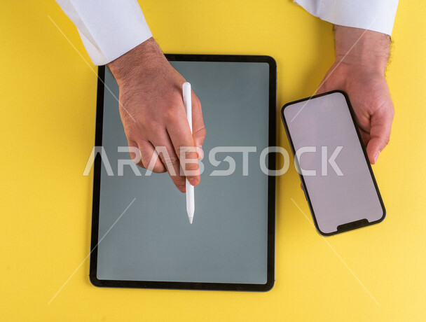 صورة مقربة ليد رجل عربي خليجي سعودي يحمل الهاتف المحمول ويستخدام جهاز التابلت ، استخدام قلم التابلت لتدوين ملاحظات خاصة بالعمل ،شرح خطوات العمل ، اعلان ترويجي لتطبيق وموقع ويب ،استخدام التقنيات الحديثة في الاعمال الادارية