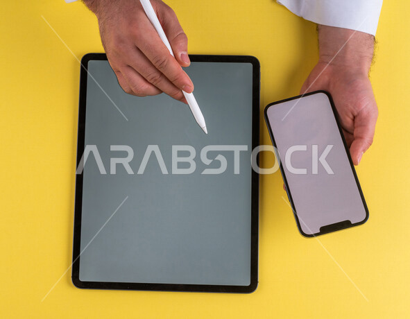 صورة مقربة ليد رجل عربي خليجي سعودي يحمل الهاتف المحمول ويستخدام جهاز التابلت ، استخدام قلم التابلت لتدوين ملاحظات خاصة بالعمل ،شرح خطوات العمل ، اعلان ترويجي لتطبيق وموقع ويب ،استخدام التقنيات الحديثة في الاعمال الادارية