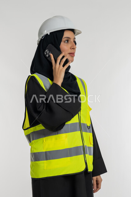 مهن ووظائف نسائية، دمج التكنولوجيا الحديثة بالهندسة، العمل في القطاع الهندسي, بورتريه من الجانب لمهندسة عربية خليجية امارتية ترتدي خوذة وسترة الحماية تقوم باجراء مكالمة هاتفية، شابة سعودية تحمل بيدها الهاتف المحمول، خلفية بيضاء