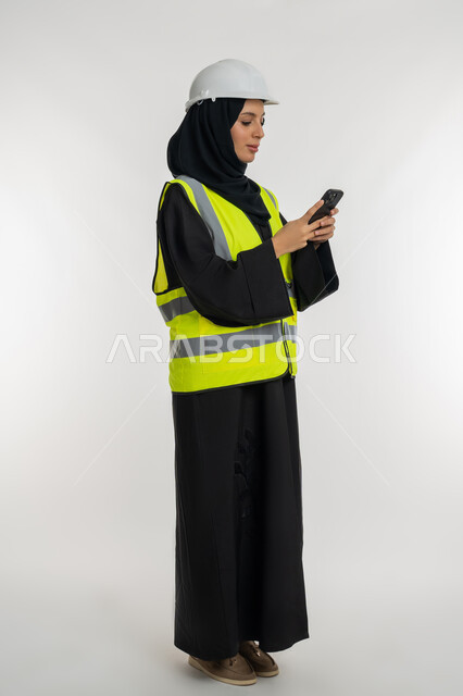 دمج التكنولوجيا الحديثة بالهندسة، مهن ووظائف نسائية، العمل في القطاع الهندسي, بورتريه من الجانب لمهندسة عربية خليجية امارتية ترتدي خوذة وسترة الحماية تقوم بمتابعة سير الاعمال عن بعد، شابة سعودية تحمل بيدها الهاتف المحمول، صورة لكامل الجسم، خلفية بيضاء