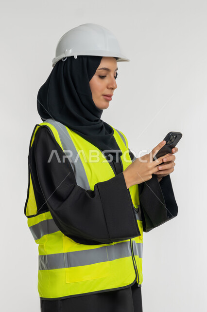 مهن ووظائف نسائية، دمج التكنولوجيا الحديثة بالهندسة، العمل في القطاع الهندسي, بورتريه من الجانب لمهندسة عربية خليجية امارتية ترتدي خوذة وسترة الحماية تقوم بمتابعة سير الاعمال عن بعد، شابة سعودية تحمل بيدها الهاتف المحمول، خلفية بيضاء