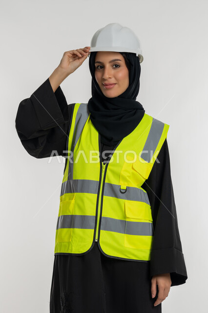 Working in the engineering sector, women's jobs and professions, the concept of engineering project management, a portrait of an Arab Gulf Emirati female architect wearing a helmet and a protective vest, touching her hat with expressions of self-confidence, a young Saudi woman looking at the camera with gestures of pleasure, white background