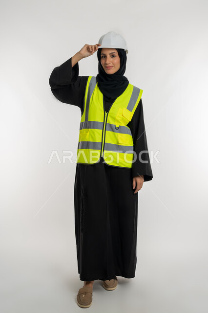 وظائف ومهن نسائية, العمل في القطاع الهندسي, مفهوم ادارة المشاريع الهندسية, بورتريه لمهندسة معمارية عربية خليجية امارتية ترتدي خوذة وسترة الحماية تمس قبعتها بتعابير الثقة بالنفس، شابة سعودية تنظر الكاميرا بايماءات السرور، صورة لكامل الجسم، خلفية بيضاء