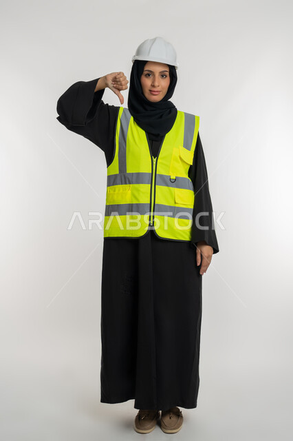 تقدم وتطور الامارات على يد أبناء الوطن، الشعور بالرفض والامتناع, مفهوم الهندسة وأعمال البناء, شابة سعودية تخفض اصبع الابهام بايماءات الانزعاج, بورتريه لمهندسة عربية خليجية اماراتية محجبة ترتدي سترة وخوذة الحماية تعمل في القطاع الهندسي, صورة لكامل الجسم، خلفية بيضاء