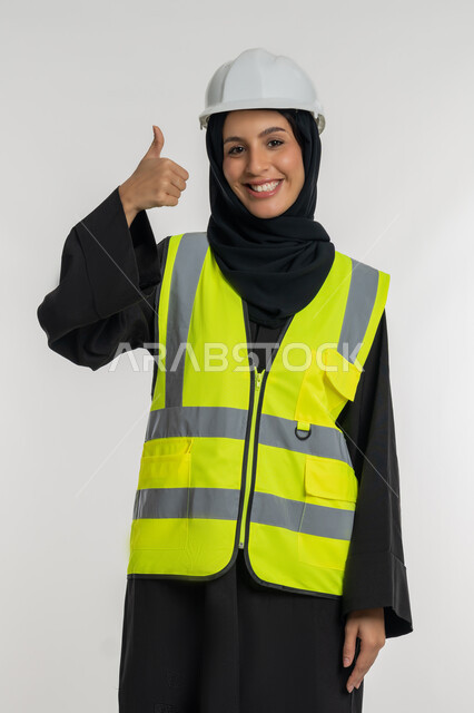 Mark of quality and excellence, progress and development of the Emirates at the hands of the sons of the nation, concept of engineering and construction work, young Saudi woman raising thumbs up in approval gestures, portrait of a veiled Arab Gulf Emirati engineer wearing a jacket and protective helmet working in the engineering sector, white background