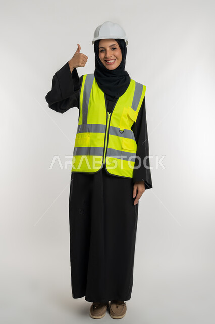 The concept of engineering and construction works, a mark of quality and excellence, the progress and development of the Emirates at the hands of the sons of the homeland, a young Saudi woman raising her thumb in a gesture of approval, a portrait of a veiled Gulf Arab Emirati engineer wearing a jacket and a protective helmet working in the engineering sector, a full body image, a white background