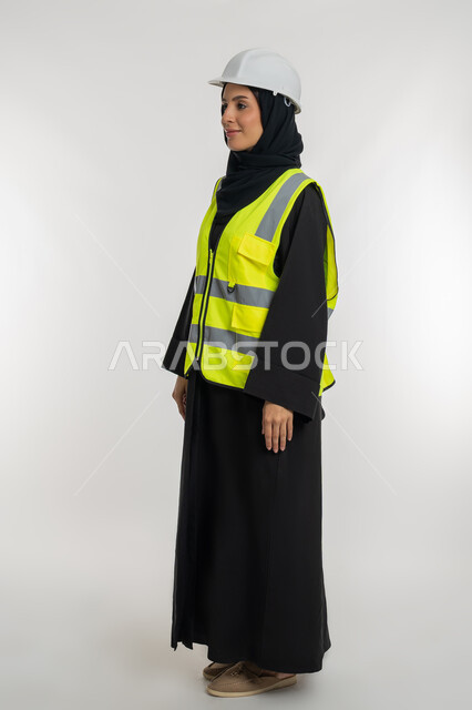 مفهوم ادارة المشاريع الهندسية,  وظائف ومهن نسائية, شابة سعودية تعمل في القطاع الهندسي, الوقوف بايماءات الثقة,  بورتريه من الجانب لمهندسة معمارية عربية خليجية امارتية ترتدي خوذة وسترة الحماية تقف باستقامة، صورة لكامل الجسم، خلفية بيضاء