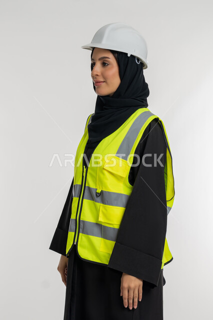 وظائف ومهن نسائية, مفهوم ادارة المشاريع الهندسية, العمل في القطاع الهندسي, الوقوف بايماءات الثقة,  بورتريه من الجانب لمهندسة معمارية عربية خليجية امارتية ترتدي خوذة وسترة الحماية، شابة سعودية تنظر لمكان ما بايماءات السرور، خلفية بيضاء