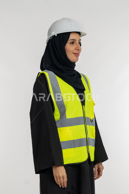 العمل في القطاع الهندسي, وظائف ومهن نسائية, مفهوم ادارة المشاريع الهندسية, الوقوف بايماءات الثقة,  بورتريه من الجانب لمهندسة معمارية عربية خليجية امارتية ترتدي خوذة وسترة الحماية، شابة سعودية تنظر لمكان ما بايماءات السرور، خلفية بيضاء