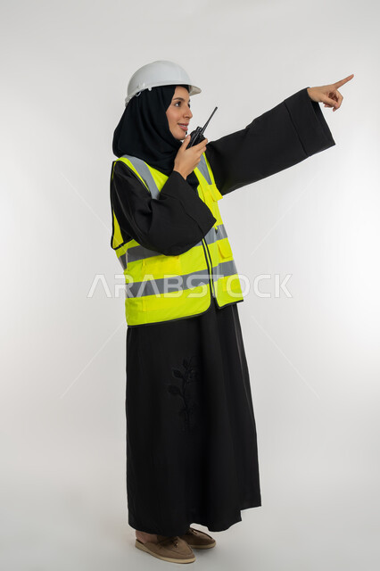 الإشراف على المشاريع، تحديد موقع شيء ما، العمل في القطاع الهندسي، بورتريه من الجانب لمهندسة عربية خليجية اماراتية ترتدي سترة وخوذة الحماية تقوم باستخدام جهاز الاتصال اللاسلكي، شابة سعودية تشير باصبع السبابة لمكان ما، صورة لكامل الجسم، خلفية بيضاء