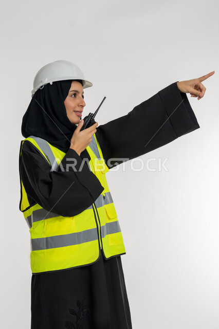 تحديد موقع شيء معين، متابعة سير الاعمال، العمل في القطاع الهندسي، الإشراف على المشاريع، بورتريه من الجانب لمهندسة عربية خليجية اماراتية ترتدي سترة وخوذة الحماية تقوم باستخدام جهاز الاتصال اللاسلكي، شابة سعودية تشير باصبع السبابة لمكان ما، خلفية بيضاء