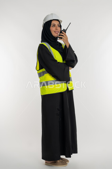 العمل في القطاع الهندسي، متابعة سير الاعمال، الإشراف على المشاريع، بورتريه من الجانب لمهندسة عربية خليجية اماراتية ترتدي سترة وخوذة الحماية تقوم باستخدام جهاز الاتصال اللاسلكي، مهن وظائف هندسية، شابة سعودية تقف بايدي كتقاطعة، صورة لكامل الجسم، خلفية بيضاء