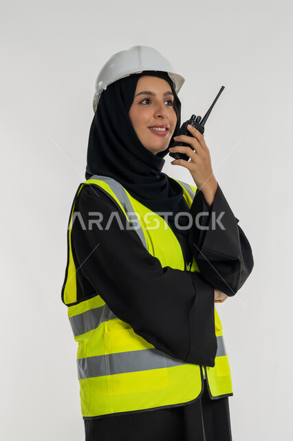 متابعة سير الاعمال، العمل في القطاع الهندسي، الإشراف على المشاريع، بورتريه من الجانب لمهندسة عربية خليجية اماراتية ترتدي سترة وخوذة الحماية تقوم باستخدام جهاز الاتصال اللاسلكي، مهن وظائف هندسية، شابة سعودية تقف بايدي كتقاطعة، خلفية بيضاء