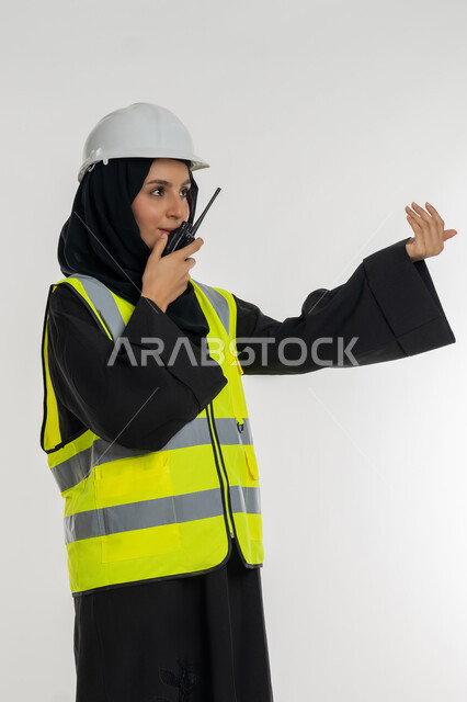 Engineering jobs, business follow-up, work in the engineering sector, project supervision, side portrait of an Arab Gulf Emirati female engineer wearing a protective jacket and helmet using a walkie-talkie, a young Saudi woman making gestures to ask to come, white background