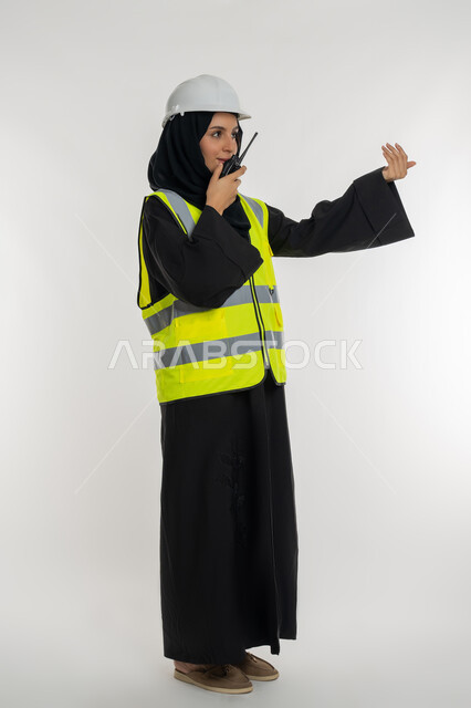 Follow up on work progress, work in engineering sector, project supervision, side portrait of an Arab Gulf Emirati female engineer wearing a protective jacket and helmet using a walkie-talkie, a young Saudi woman gestures with her hand asking to come, full body photo, white background