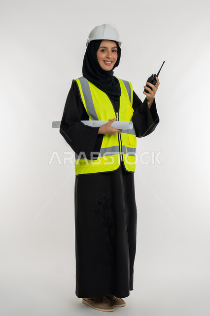 الإشراف على المشاريع، ايماءات السرور والسعادة، العمل في القطاع الهندسي، بورتريه لمهندسة عربية خليجية اماراتية ترتدي سترة وخوذة الحماية تقوم باستخدام جهاز الاتصال اللاسلكي، شابة سعودية تحمل بيدها مخطط المشروع، صورة لكامل الجسم، خلفية بيضاء
