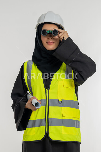 Love of adventure and exploration, looking forward to future goals, working in the engineering sector, construction plans and projects, portrait of a smiling veiled Saudi Arabian Gulf woman holding a project plan in her hand, a young Saudi woman looking through binoculars, white background