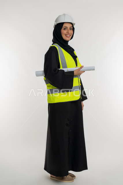 العمل في القطاع الهندسي، تعابير السرور والسعادة، مخططات و مشاريع اعمارية، مهن ووظائف نسائية هندسية، بورتريه من الجانب لامرأة عربية خليجية سعودية محجبة مبتسمة تحمل بيدها مخطط المشروع، شابة سعودية تقوم بالاشراف على العمل، صورة لكامل الجسم، خلفية بيضاء