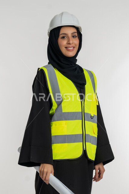 النظر إلى الكاميرا بتعابير السرور والسعادة، العمل في القطاع الهندسي، مخططات و مشاريع اعمارية، مهن ووظائف نسائية هندسية، بورتريه لامرأة عربية خليجية سعودية محجبة مبتسمة تحمل بيدها مخطط المشروع، شابة سعودية تقوم بالاشراف على العمل، خلفية بيضاء