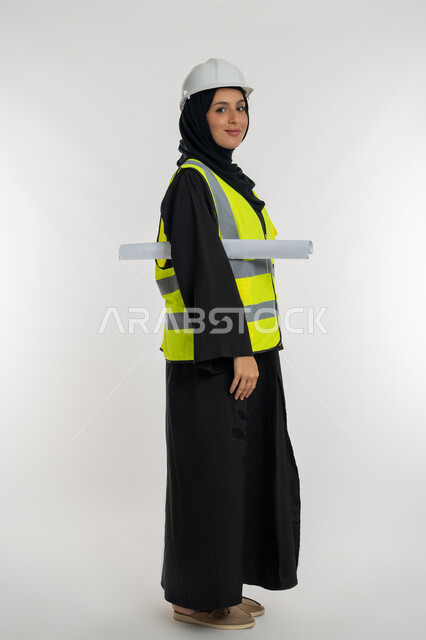 العمل في القطاع الهندسي، تعابير السرور والسعادة، مخططات و مشاريع اعمارية، مهن ووظائف نسائية هندسية، بورتريه من الجانب لامرأة عربية خليجية سعودية محجبة مبتسمة تحمل بيدها مخطط المشروع، شابة سعودية تقوم بالاشراف على العمل، صورة لكامل الجسم، خلفية بيضاء
