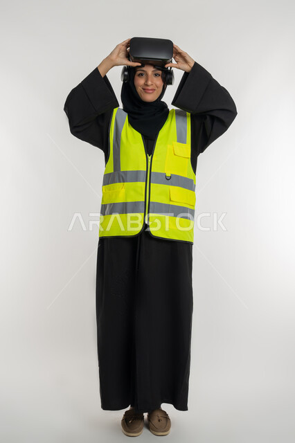 الاندماج مع الهندسة الافتراضية، تطوير المجالات الهندسية عبر نظارات الفي آر، رؤية مستقبلية 2030 ، بورتريه لمهندسة عربية خليجية اماراتية ترتدي سترة وخوذة الحماية تضع نظارة الواقع الافتراضي، شابة سعودية تنظر الى الكاميرا بايماءات السرور، صورة لكامل الجسم، خلفية بيضاء