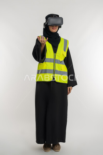 تطوير المجالات الهندسية عبر نظارات الفي آر، التفاعل مع الهندسة الافتراضية، بورتريه لمهندسة عربية خليجية اماراتية ترتدي سترة وخوذة الحماية تضع نظارة الواقع الافتراضي، شابة سعودية ترفع يدها بايماءات حمل عنصر ما، صورة لكامل الجسم، خلفية بيضاء