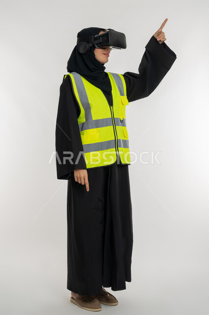 Interacting with virtual engineering, locating something, developing engineering fields through VR glasses, portrait of an Arab Gulf Emirati engineer wearing a protective jacket and helmet, putting on virtual reality glasses, a young Saudi woman pointing with her index finger to a place, white background