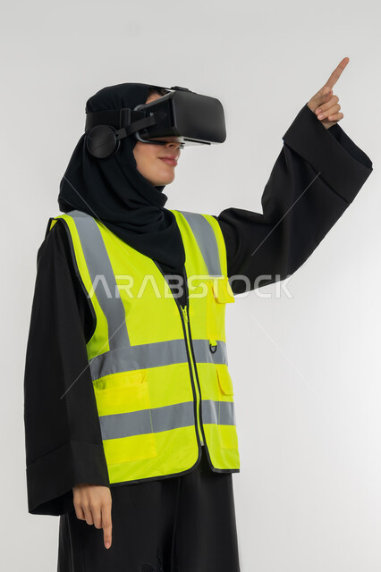 التفاعل مع الهندسة الافتراضية، تحديد موقع شيء ما، تطوير المجالات الهندسية عبر نظارات الفي آر، بورتريه لمهندسة عربية خليجية اماراتية ترتدي سترة وخوذة الحماية تضع نظارة الواقع الافتراضي، شابة سعودية تشير باصبع السبابة لمكان ما، خلفية بيضاء