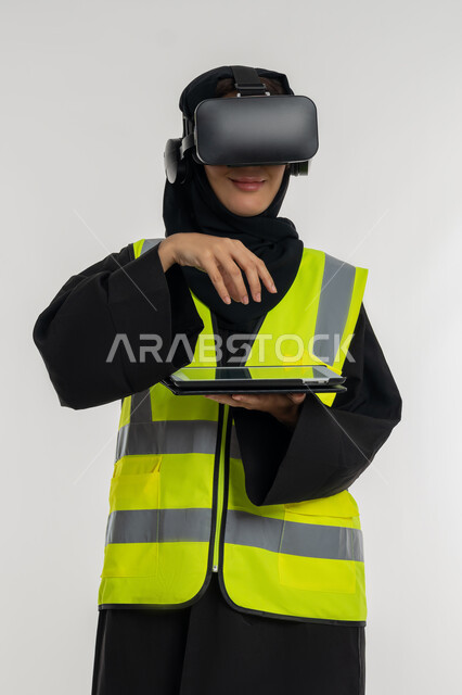 Interacting with virtual engineering, developing engineering fields through VR glasses, portrait of an Arab Gulf Emirati female engineer wearing a protective jacket and helmet, wearing virtual reality glasses and holding a tablet, a young Saudi woman raising her hands with gestures of integration, white background