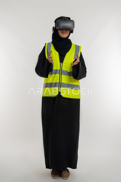 Expressions of holding an item, interacting with virtual engineering, developing engineering fields through VR glasses, portrait of an Arab Gulf Emirati female engineer wearing a protective jacket and helmet, putting on virtual reality glasses, a young Saudi woman raising her hands with scaling gestures, full body image, white background