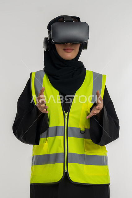 تعابير حمل عنصر ما، التفاعل مع الهندسة الافتراضية، رؤية مستقبلية 2030، تطوير المجالات الهندسية عبر نظارات الفي آر، بورتريه مقرب لمهندسة عربية خليجية اماراتية ترتدي سترة وخوذة الحماية تضع نظارة الواقع الافتراضي، شابة سعودية ترفع يديها بايماءات التحجيم، خلفية بيضاء