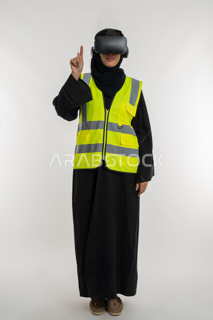 تحديد موقع شيء ما، تطوير المجالات الهندسية عبر نظارات الفي آر، الاندماج مع الهندسة الافتراضية، بورتريه لمهندسة عربية خليجية اماراتية ترتدي سترة وخوذة الحماية تضع نظارة الواقع الافتراضي، شابة سعودية تشير باصبع السبابة للاعلى، صورة لكامل الجسم، خلفية بيضاء