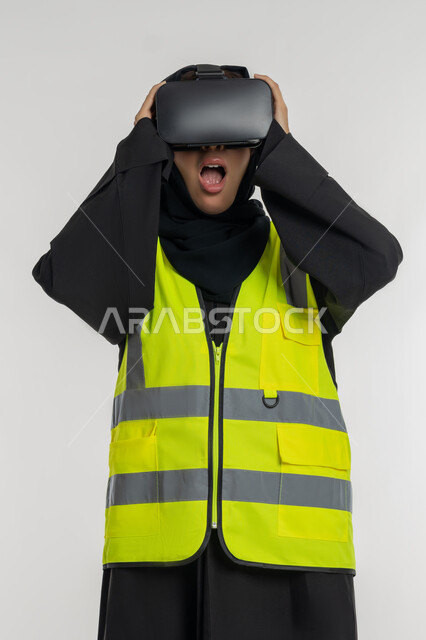 Expressions of shock and surprise, development of engineering fields through VR glasses, future vision 2030, integration with virtual engineering, portrait of an Arab Gulf Emirati female engineer wearing a protective jacket and helmet, putting on virtual reality glasses, a young Saudi woman holding glasses with gestures of astonishment, white background