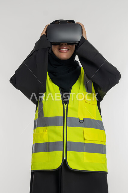 تطوير المجالات الهندسية عبر نظارات الفي آر، رؤية مستقبلية 2030 ، الاندماج مع الهندسة الافتراضية، بورتريه لمهندسة عربية خليجية اماراتية ترتدي سترة وخوذة الحماية تضع نظارة الواقع الافتراضي، شابة سعودية تمسك النظارات بايماءات السرور، خلفية بيضاء