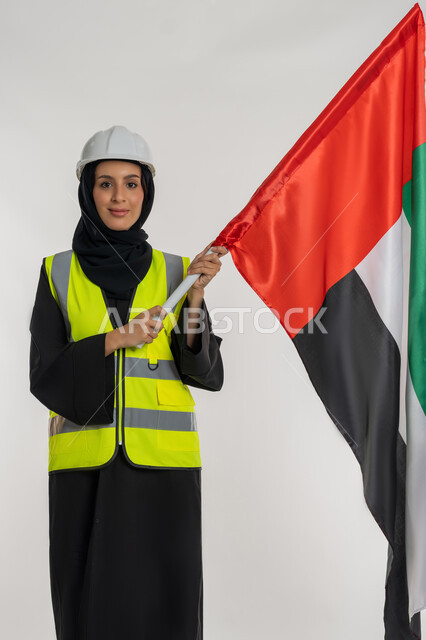 ايماءات الخر والاعتزاز بالانتماء للوطن، الاحتفال بيوم العلم الاماراتي 3 نوفمبر، ذكرى يوم الاستقلال 2 ديسمبر، بورتريه لمهندسة عربية خليجية اماراتية ترتدي سترة وخوذة الحماية تحمل بيدها علم دولة الإمارات المتحدة، خلفية بيضاء