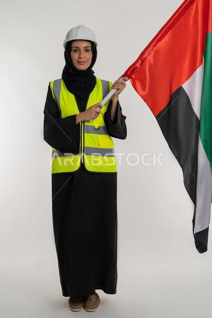 ايماءات الخر والاعتزاز بالانتماء للوطن، الاحتفال بيوم العلم الاماراتي 3 نوفمبر، ذكرى يوم الاستقلال 2 ديسمبر، بورتريه لمهندسة عربية خليجية اماراتية ترتدي سترة وخوذة الحماية تحمل بيدها علم دولة الإمارات المتحدة، صورة لكامل الجسم، خلفية بيضاء