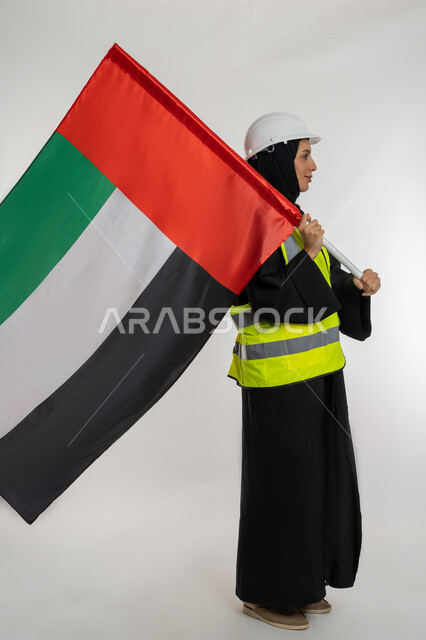 ذكرى يوم الاستقلال 2 ديسمبر، بورتريه من الجانب لمهندسة عربية خليجية اماراتية ترتدي سترة وخوذة الحماية تحمل بيدها علم دولة الإمارات المتحدة، إيماءات الفخر والإعتزاز بالانتماء للوطن، الاحتفال بيوم العلم الإماراتي 3 نوفمبر، صورة لكامل الجسم، خلفية بيضاء