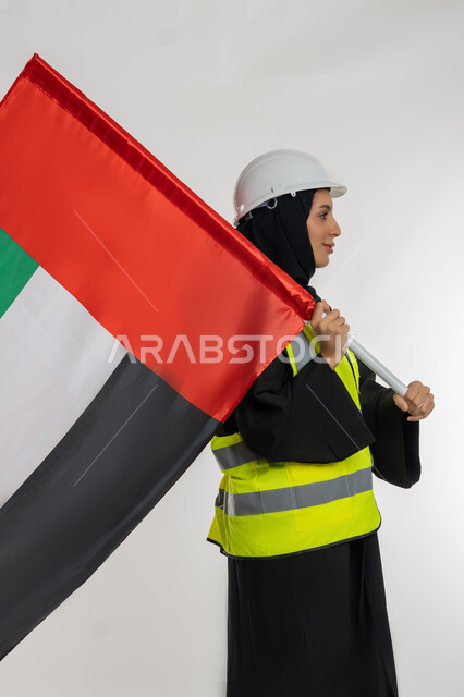 الاحتفال بيوم العلم الاماراتي 3 نوفمبر، ذكرى يوم الاستقلال 2 ديسمبر، بورتريه من الجانب لمهندسة عربية خليجية اماراتية ترتدي سترة وخوذة الحماية تحمل بيدها علم دولة الإمارات المتحدة، إيماءات الفخر والإعتزاز بالانتماء للوطن، خلفية بيضاء