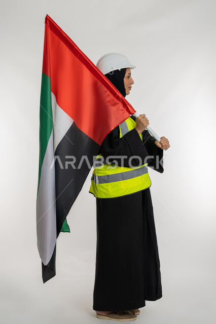 ذكرى يوم الاستقلال 2 ديسمبر، بورتريه من الجانب لمهندسة عربية خليجية اماراتية ترتدي سترة وخوذة الحماية تحمل بيدها علم دولة الإمارات المتحدة، إيماءات الفخر والإعتزاز بالانتماء للوطن، الاحتفال بيوم العلم الإماراتي 3 نوفمبر، صورة لكامل الجسم، خلفية بيضاء